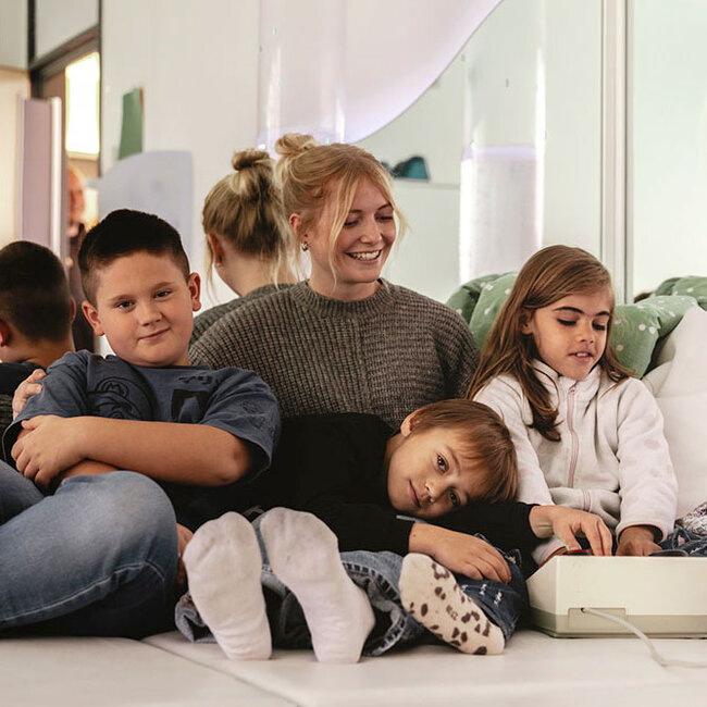 zwei Jungen und ein Mädchen mit einer Betreuerin ruhen im Snoezelraum sitzend