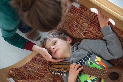 Jugendlicher mit Taubblindheit liegt in einer Klangwiege, die Betreuerin legt die Hand an seinen Kopf und beugt sich über ihn