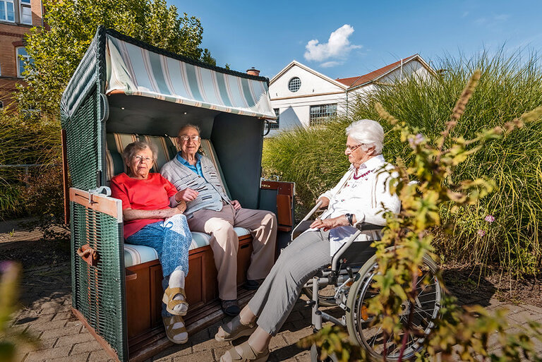 Drei ältere Menschen sitzen im Garten - ein Mann und eine Dame im Strandkorb, eine Dame im Rollstuhl