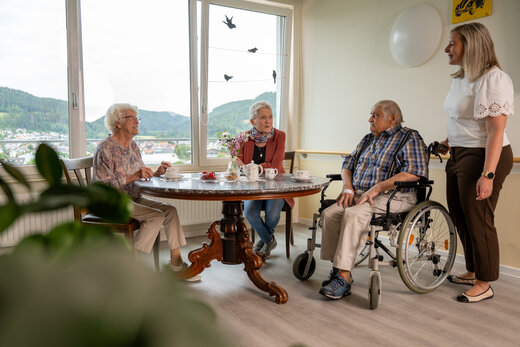 Aufenthaltsbereich einer Wohngruppe - zwei Frauen und ein Mann im Rollstuhl sitzen am Kaffeetisch eine weitere Frau steht nebendran