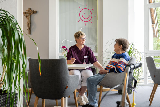 Pflegeperson und betreute ältere Dame unterhalten sich in einer Sitzecke