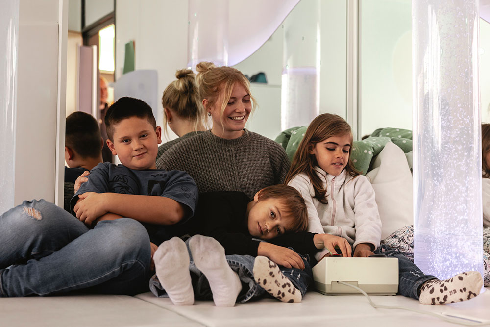 zwei Jungen und ein Mädchen mit einer Betreuerin ruhen im Snoezelraum sitzend